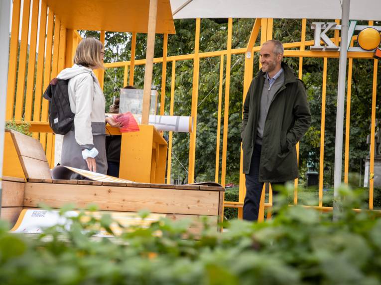 Zwei Personen in einem Gespräch in einem Kiosk aus orangefarbenen Brettern.